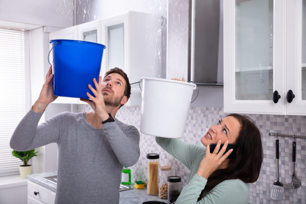 Worried Young Woman Calling Plumber On Cellphone While Man Collecting Leakage Water From Ceiling